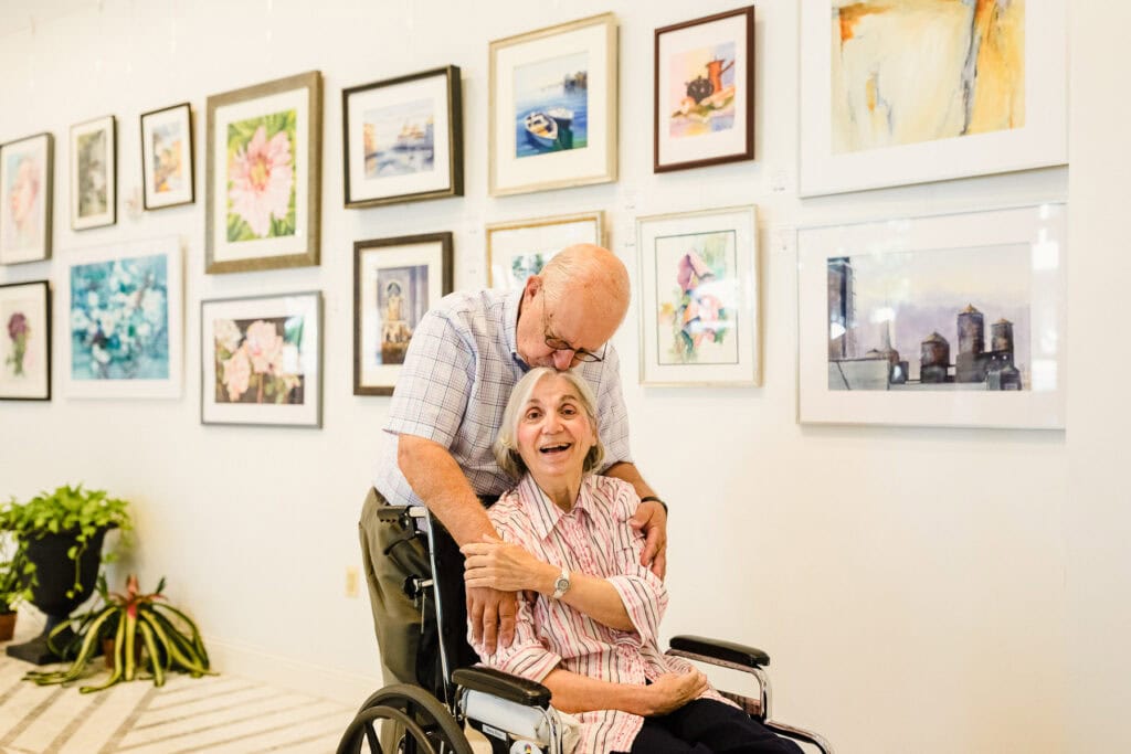 older man kissing older woman in wheelchair on the head